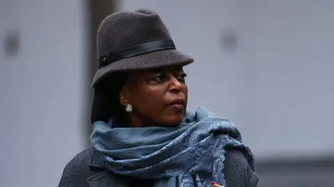 Headshot of Diezani Alison-Madueke looking off-camera wearing a grey brimmed hat with a blue scarf and grey coat.