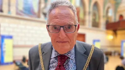 Dennis Meredith with short grey hair wearing glasses and a grey jacket with a black and white spotted shirt and red tie. He is wearing a gold chain of office over his shoulders. He is standing in Northampton Guidhall.