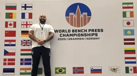 Ram Patten Ram Patten stood infront of a walk which says 'world bench press championships', surrounded by world flags.