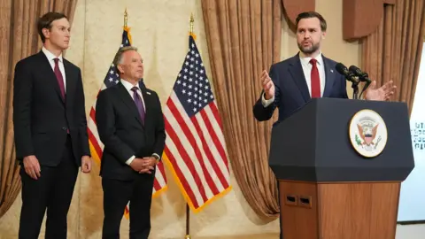 US Vice-President JD Vance stands at a podium with Jared Kushner and Steve Witkoff behind him.