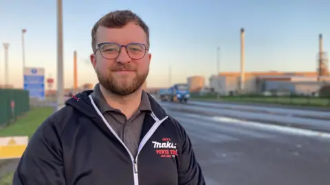 The photo shows Tom Smith smiling at the camera. He has brown hair and glasses. He is wearing a black zip jacket with a grey polo top underneath. He is standing in front of a sunrise, which is behind the Steelworks. There are several towers behind him, and a road to the right hand side. 