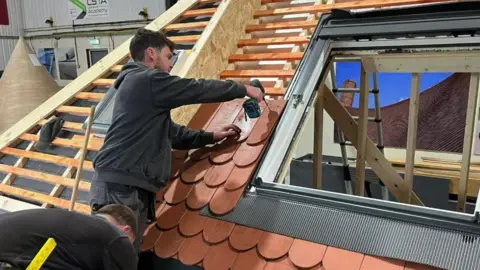 Joe holds a drills over some adobe-coloured tiles next to a window while working on a mock-up roof rig.