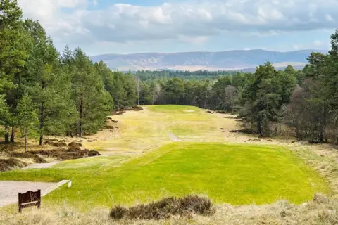 Green Grantown-on-Spey golf hole, lined by trees, with hills in the distance, and a cloudy blue sky.