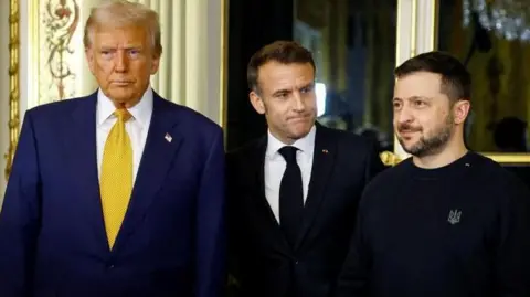 Trump, Macron and Zelensky standing next to each other during a meeting at the French Elysee Palace. Trump and Macron wear suits, while Zelensky is more casually dressed in a black sweatshirt.