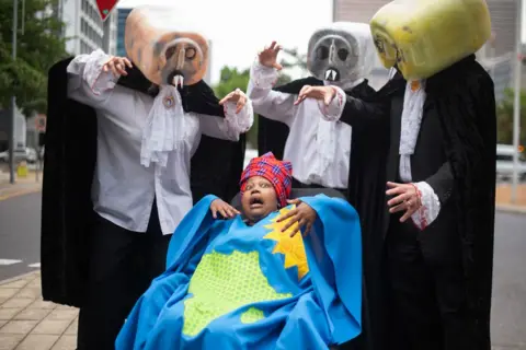 RODGER BOSCH / AFP A protestor from Extinction Rebellion Cape Town (C), wearing clothes that represents Africa, is being threatened by 'Fossil Fuel Vampires' dressed in lawyers outfits during a demonstration against the ongoing use of fossil fuels, outside the venue where the Africa Oil Week Conference is being held.