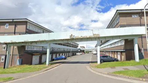 LDRS The picture show the current estate which has a tatty concrete entrance to to rows of maisonettes behind with cars parked outside some