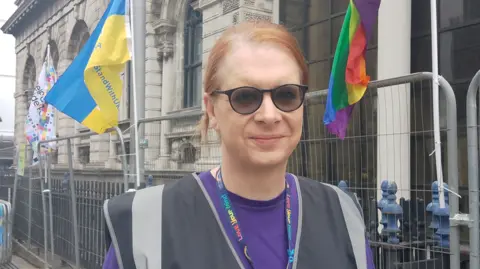 Kirsty smiling at the camera. In the background temporary fencing and pride flags