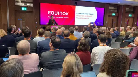 BBC A busy room with a woman at the front, presenting. Behind her is a big screen with the words EQUINOX written on them,