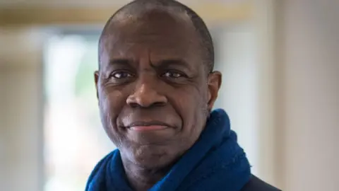 Clive Myrie Clive Myrie smiling at the camera. He is a black man with short back hair and he is wearing blue scarf.