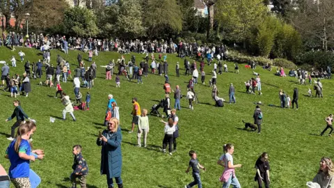 Large crowds of people of all ages are scattered over the large grassy slope with a line of families watching from the top ridge with trees behind.