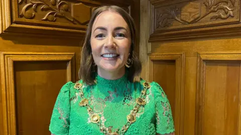Grace Uí Niallais stands in front of wooden panelling. She is smiling and wearing a green dress. She has shoulder length brown hair and is wearing a mayoral chain around her neck