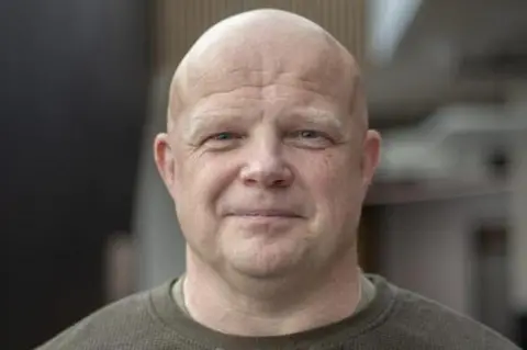 Andrew Bartlett Andrew Bartlett in the centre of the photograph smiling at the camera. He is middle aged, bald and has blue eyes. He wears a khaki long-sleeve t-shirt. 