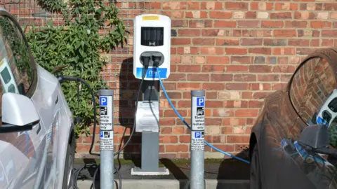 East Riding of Yorkshire Council Two electric vehicle charging points are plugged in at a white stand next to a grey and black car. Two silver posts say parking on them.