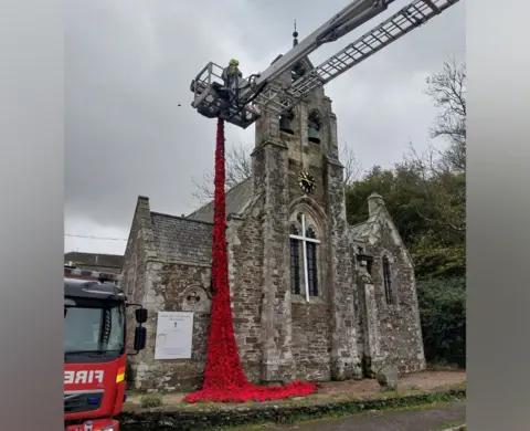 Kirsten Lean A firefighter in a cherry picker is seen draping the red poppy display on to a stone church building. Part of a fire engine is to the left of the image.