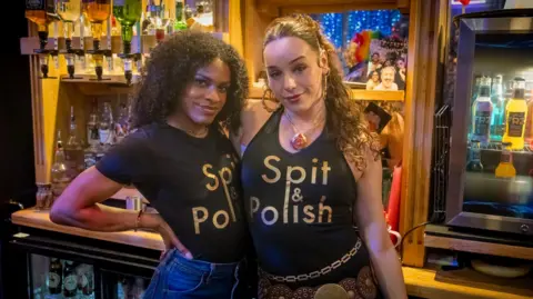 Channel 4 Shakeel is a black woman with curly black long hair and Iz is a white woman with long curly brown hair. They both wear black t-shirt with letters in gold saying Spit & Polish and stand close together