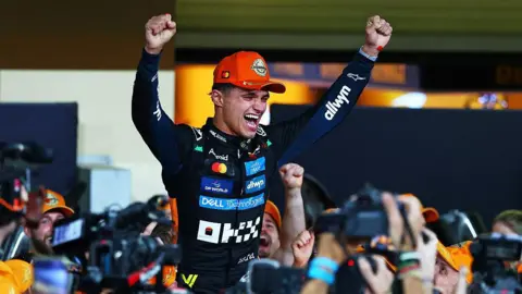 Getty Images A racing driver dressed in a black racing suit with sponsors' badges all over the front. He has his hands aloft and is being supported on the shoulders of those in crowd. 