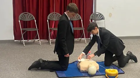 Two male students using a defibrillator on a practice dummy. They are wearing black school uniforms. The dummy is on a blue mat. The boy to the right is placing defibrillator receptors on the dummy.