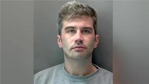 Kieran Roche: a police custody image of a man with short light-brown hair wearing a grey round-necked T-shirt