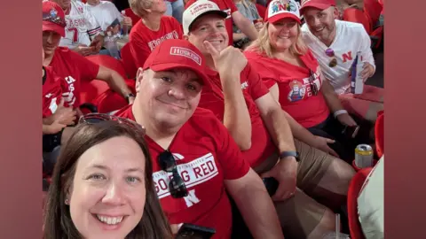 Contributed A woman with long brown hair and sunglasses on her head takes a selfie with friends who sit in the stands during a sport event. They all wear similar red tops as well as red and white caps. 