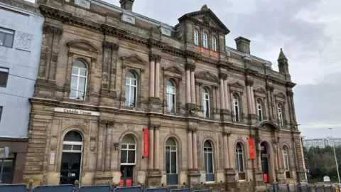 Large impressive Victorian light stone, three storey building in the centre of the image. The building has for visible red banners attached to two of its doors and partway up the wall by its second and fifth window frames.
In the foreground are a series nine square, solid blue metal protective barriers, transitioning into a series of see through blue barriers, a demarcation for trams. 