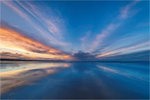 Alan Butterfield Un cielo azul y naranja reflejado en el agua en una playa.