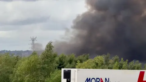 A photo showing plumes of black smoke behind a row of trees and a lorry.