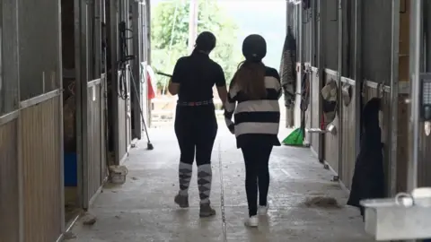 Two young girls wearing riding hats and with their backs to the camera walk between two rows of stables. 