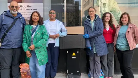 Six people stood together, smiling outside a shop window. Ladies in centre propping up an insulated food box. 