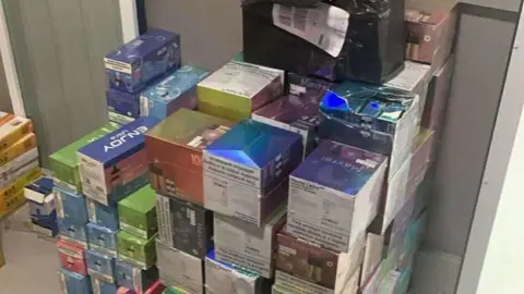 About 30-40 different coloured cardboard boxes storing illegal vapes stacked one on top of the other in a storage unit. Different vape brand names are on the boxes.