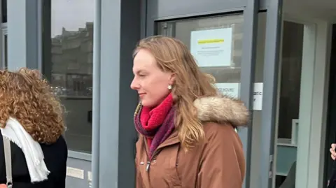 BBC Dr Beth Upton - a transgender woman with long blonde hair, a red scarf and a brown jacket - leaves a building