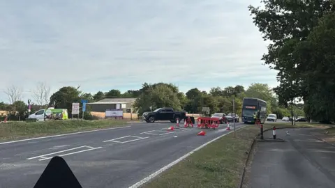 Luke Deal/BBC Vehículos girando en una carretera cerrada. Hay barreras en la calzada. Detrás, un autobús, coches y furgonetas giran hacia otra carretera. Más allá se ven campos y árboles.