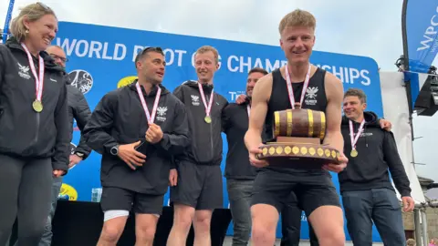 A rowing crew celebrate winning the World Pilot Gig Championships on a stage in the Isles of Scilly. The team is from Falmouth and are all wearing black clothing with the club's emblem on it. A man with short blonde hair is holding the trophy which has a mini wooden barrel on it.