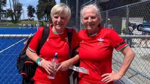 Twin sisters both aged 66, with silver hair, wearing their welsh jersey shirts outside on a playing field. 