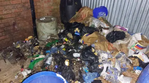 Rubbish can be seen built up on the floor outside the premises next to a bin and metal drums.