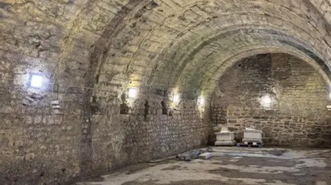 A dark underground vault has stone walls and a curved ceiling. It is illuminated with bright wall lights. The floor and walls looks damp.