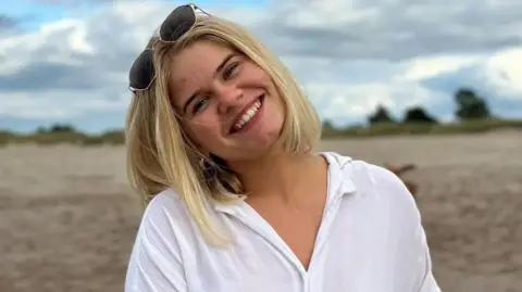 Phoenix Spencer-Horn smiling directly at the camera. She has her head tilted to the right and has blonde hair, which drapes over her shoulder and sunglasses on top of her head. She is wearing a white shirt and is standing in front of a beach, trees and blue sky which blurred in the background.