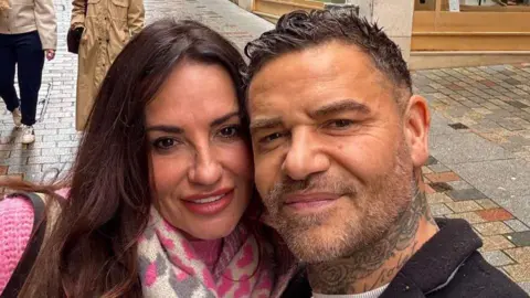 Jayne Nicholson, with long brown hair and wearing a pink jumper, stands beside George Goodayle, with a neck tattoo and wearing a black jacket