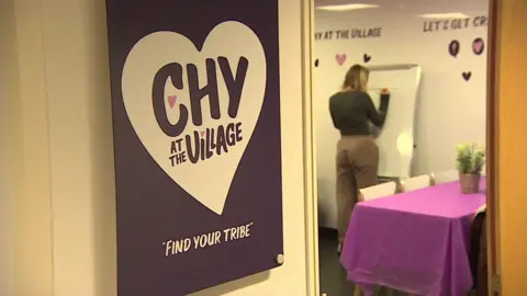 A purple sign reads CHY AT THE VILLAGE and FIND YOUR TRIBE. A woman with her back to the camera can be seen inside the hub writing on a whiteboard.