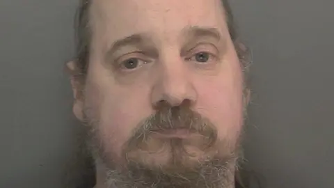 A police mugshot of Alexander Sherwood. He has long greying hair with a moustache and beard.