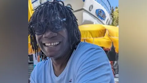 Derek Thomas is smiling at the camera with almost shoulder-length hair in dreadlocks. He is wearing sunglasses and wears a white t-shirt, sitting outside. Behind him are yellow umbrellas and the bow of a boat.