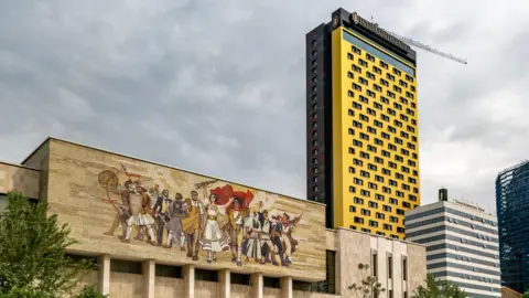 Getty Images Tirana's central Skenderbeg Square. In the foreground is a large mural from Albania's communist era in the socialist-realist style. Behind this building there are cranes and skyscrapers under construction. 