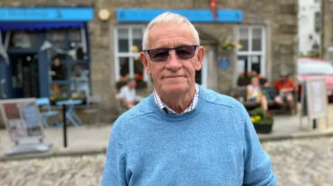 Mark Ansell/BBC A man in sunglasses and a blue jumper stands in front of stone buildings in Grassington.