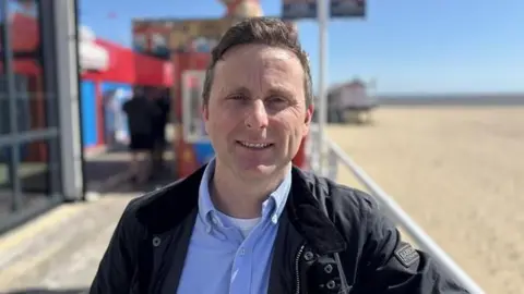 Luke Deal/BBC Joseph Abbott wears a blue shirt, open at the neck, and a navy wax jacket. He has swept back fair hair and is smiling. He is standing on the pier with the beach and sea in the background.