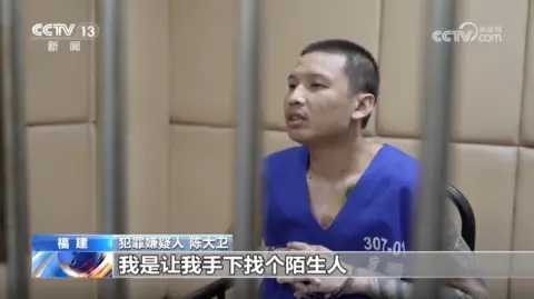 CCTV Chen Dawei wearing a blue prison vest, with his wrists in handcuffs and sitting on a chair behind bars. There are Chinese subtitles at the bottom of the screen and the CCTV logo on top corners.