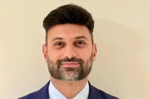 A square-jawed man smiles into the camera. He has black hair and is sporting a quiff. His closely cropped beard is black and grey. He is wearing a dark blue jacket and a light blue shirt
