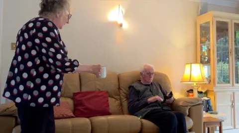 A woman with grey hair and glasses wearing a spotty blouse handing a cup of tea to a man with grey hair and glasses, who is sitting on a sofa using his mobile phone, with a table and a lamp next to him