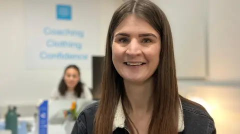 Jaz smiles into the camera. She was long brown hair and is wearing a black denim jacket with a fir lapel. Behind her, a women is blurred out sitting at a desk.