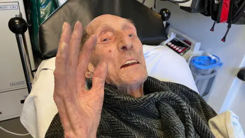 An elderly man lying on an ambulance stretcher waves at the camera.
