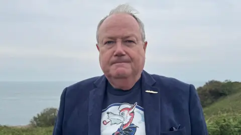 A man wearing a navy blazer over a shirt with a fox on it standing in front of grassy headlands and the sea in the background.