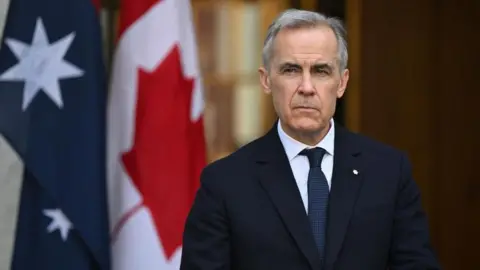 An image of Mark Carney taken straight ahead as he looks off camera. Behind him are the flag of Australia and Canada side-by-side. He is wearing a black suit and a dark blue patterned tie. He has short black and white hair that is parted on the right.